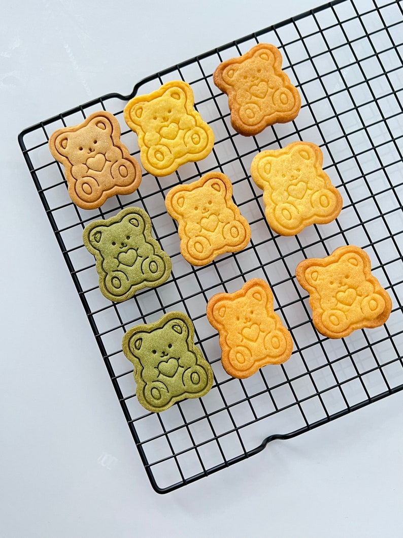 May include: Nine bear-shaped cookies on a black wire cooling rack. The cookies are in three colors: yellow, green, and a light brown. Each cookie has a heart design in the center.