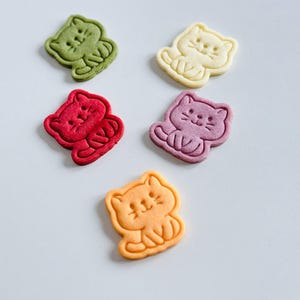 Puede incluir: Cinco galletas con forma de gato en varios colores: verde, amarillo, rojo, morado y naranja. Cada galleta tiene un contorno detallado y rasgos faciales, sobre una superficie blanca. Un dulce divertido y colorido.