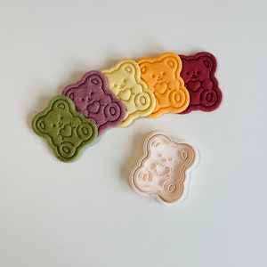 May include: Five colorful bear-shaped cookies with a heart design and a white plastic bear-shaped cookie cutter.