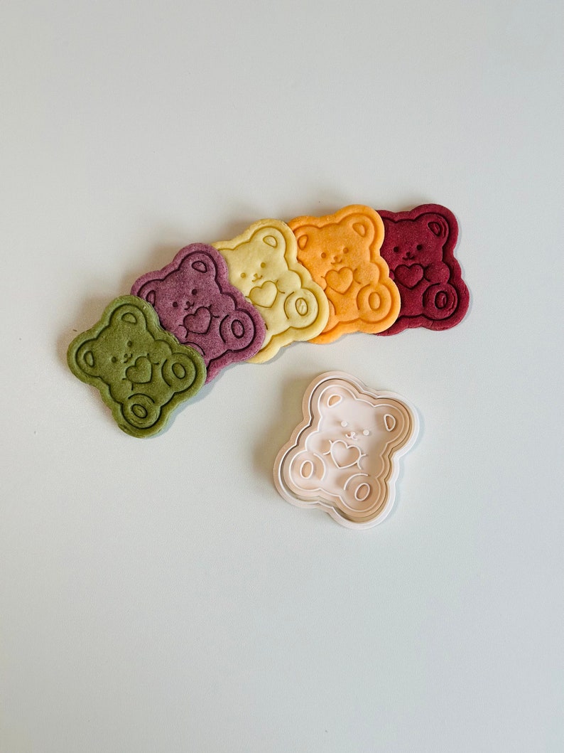 May include: Five colorful bear-shaped cookies with a heart design and a white plastic bear-shaped cookie cutter.