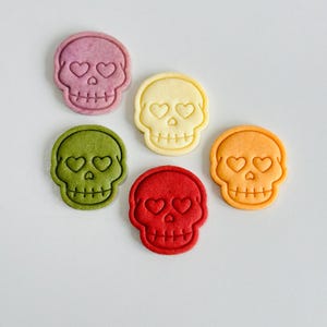 May include: Six colorful skull-shaped cookies with heart-shaped eyes. The cookies are in shades of purple, yellow, green, red, and orange. The cookies are arranged on a white surface.