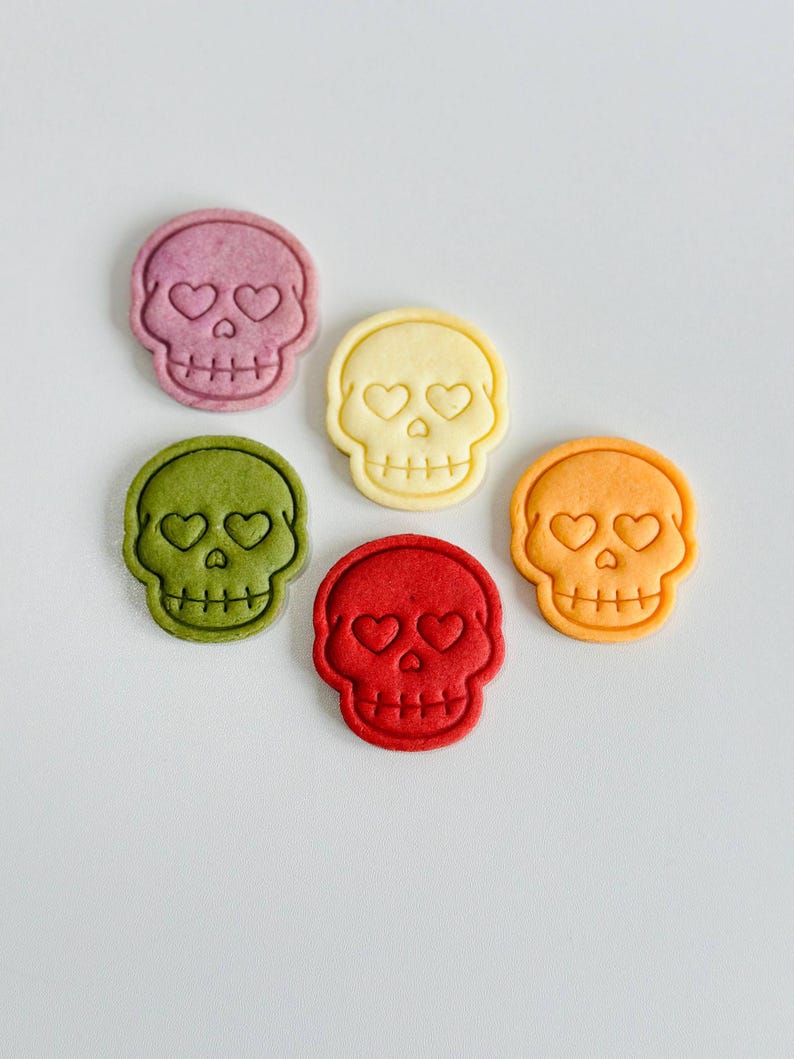 May include: Six colorful skull-shaped cookies with heart-shaped eyes. The cookies are in shades of purple, yellow, green, red, and orange. The cookies are arranged on a white surface.
