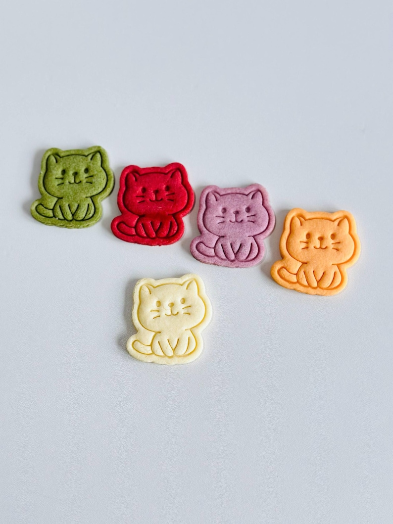 Puede incluir: Seis galletas con forma de gato en varios colores: verde, rojo, morado, naranja y amarillo. Cada galleta tiene un dise&ntilde;o sencillo, tipo caricatura, con una cara sonriente y una pose sentada. Las galletas est&aacute;n dispuestas sobre una superficie blanca.