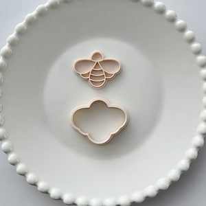 May include: Two plastic cookie cutters on a white plate. One cutter is shaped like a bee and the other is shaped like a cloud.