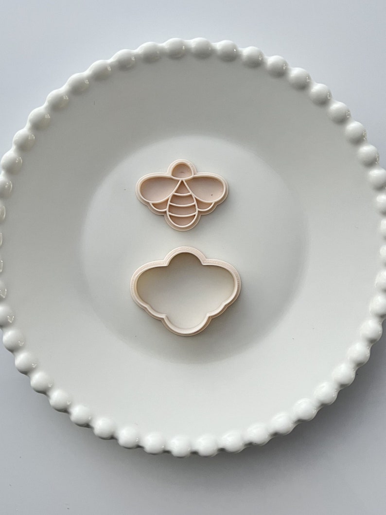 May include: Two plastic cookie cutters on a white plate. One cutter is shaped like a bee and the other is shaped like a cloud.