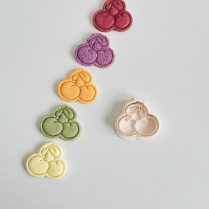 May include: Five cookies shaped like a cluster of three cherries, in various colors: red, purple, yellow, green, and light yellow. A white plastic cookie cutter shaped like a cluster of three cherries is also shown.