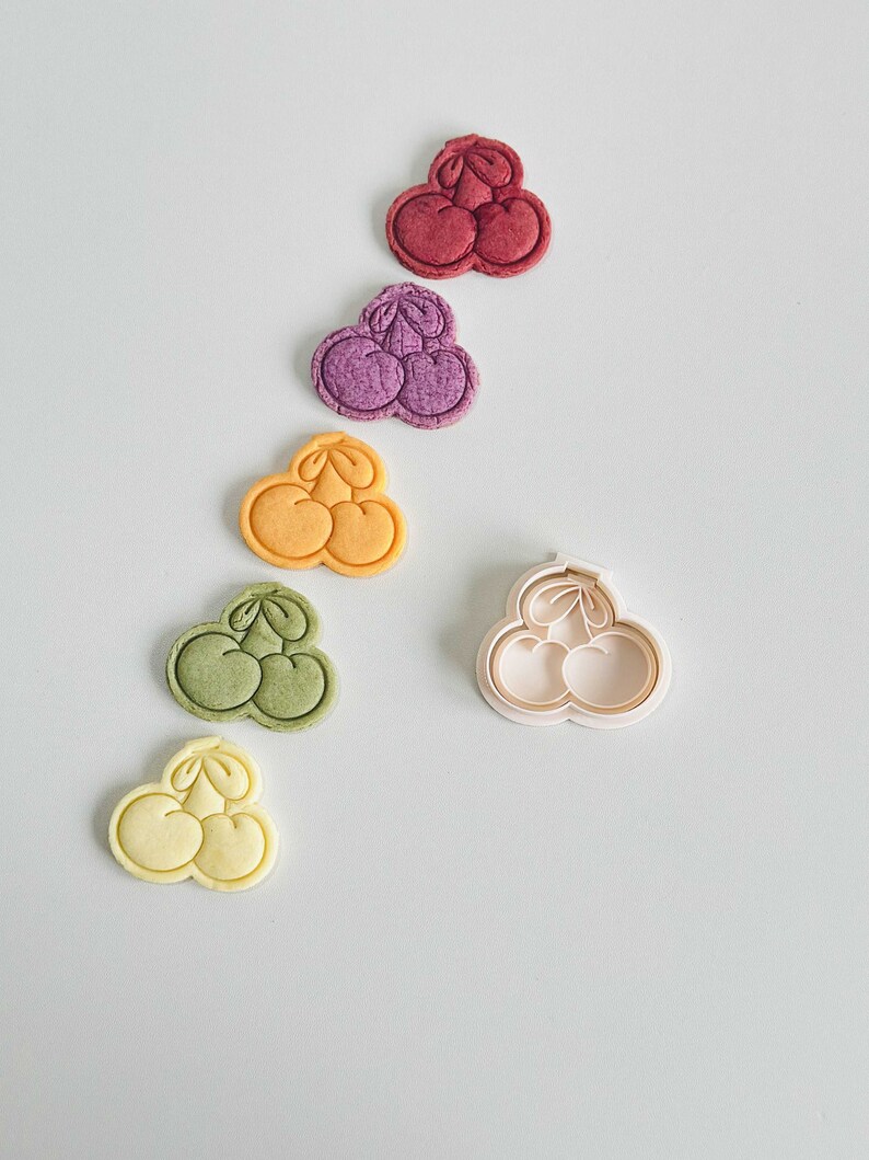 May include: Five cookies shaped like a cluster of three cherries, in various colors: red, purple, yellow, green, and light yellow. A white plastic cookie cutter shaped like a cluster of three cherries is also shown.