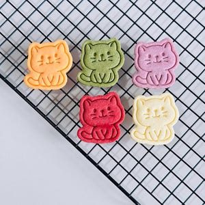 Puede incluir: Seis galletas con forma de gato en varios colores: naranja, verde, rosa, rojo y amarillo. Cada galleta tiene una cara sonriente y est&aacute; sentada. Las galletas est&aacute;n dispuestas sobre una rejilla de enfriamiento negra.
