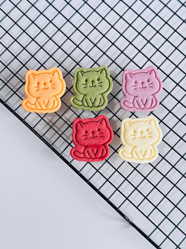Puede incluir: Seis galletas con forma de gato en varios colores: naranja, verde, rosa, rojo y amarillo. Cada galleta tiene una cara sonriente y est&aacute; sentada. Las galletas est&aacute;n dispuestas sobre una rejilla de enfriamiento negra.