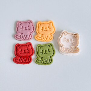 Puede incluir: Una colecci&oacute;n de galletas con forma de gato en varios colores: rosa, naranja, rojo y verde. Tambi&eacute;n hay un cortador de galletas rosa con forma de gato. Las galletas tienen rasgos faciales detallados y est&aacute;n sentadas.