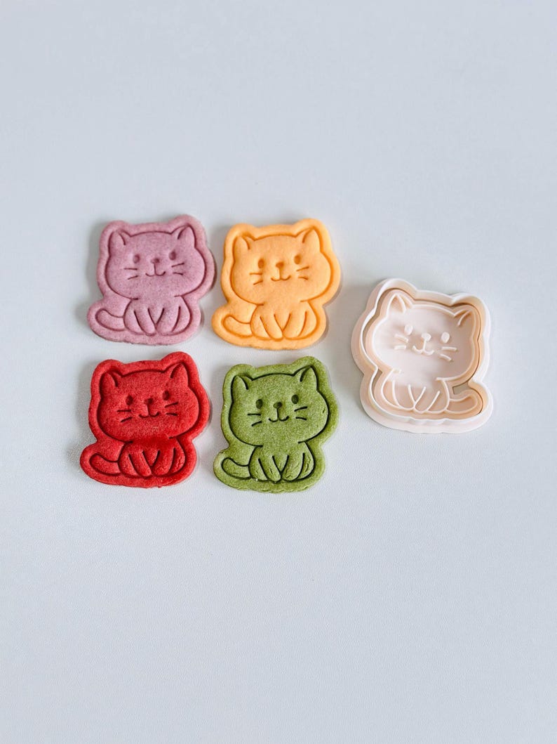 Puede incluir: Una colecci&oacute;n de galletas con forma de gato en varios colores: rosa, naranja, rojo y verde. Tambi&eacute;n hay un cortador de galletas rosa con forma de gato. Las galletas tienen rasgos faciales detallados y est&aacute;n sentadas.