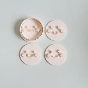 May include: A set of four light pink cookie cutters with different smiley face designs. One cutter is round with a circular rim, and the other three are individual smiley face shapes. The background is a light blue-green color.