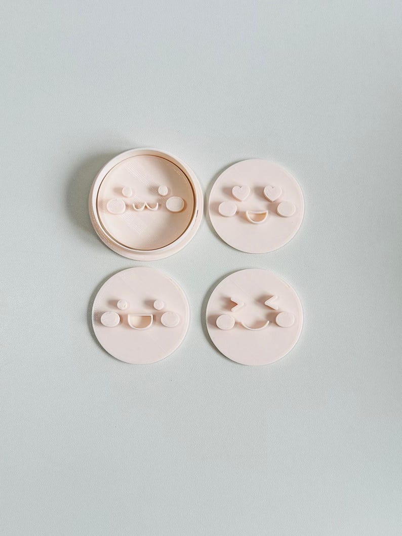 May include: A set of four light pink cookie cutters with different smiley face designs. One cutter is round with a circular rim, and the other three are individual smiley face shapes. The background is a light blue-green color.