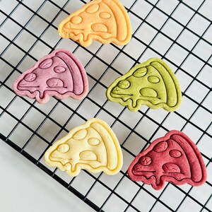 May include: Five colorful pizza-shaped cookies on a black wire rack. The cookies are yellow, pink, green, white, and red.