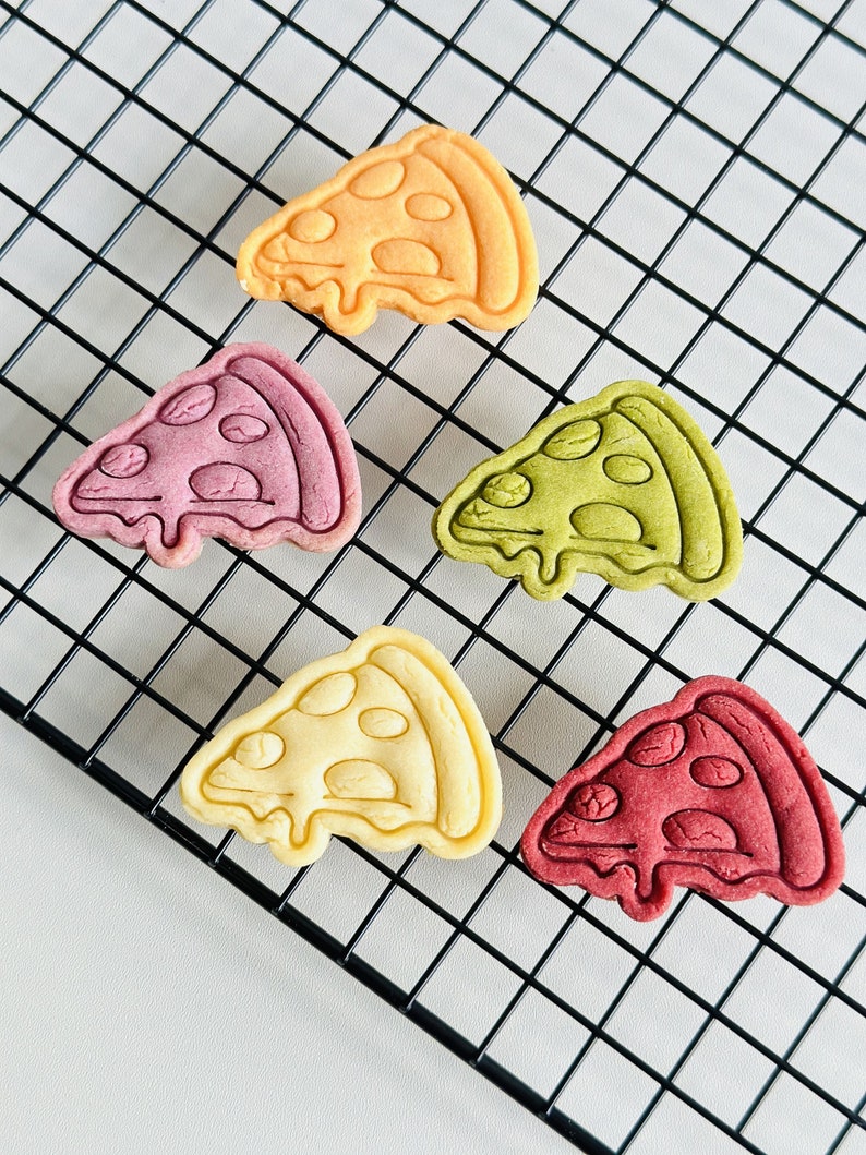 May include: Five colorful pizza-shaped cookies on a black wire rack. The cookies are yellow, pink, green, white, and red.