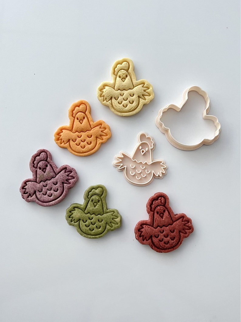 May include: Six colorful cookie cutters in the shape of a chicken. The cutters are arranged in a circle on a white surface. One cutter is a solid shape, while the other five are detailed with a chicken's face, wings, and feathers.