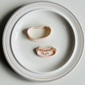 May include: Two pink plastic cookie cutters on a white plate. One cutter is shaped like a hot dog with a smiling face and the other is shaped like a bun.