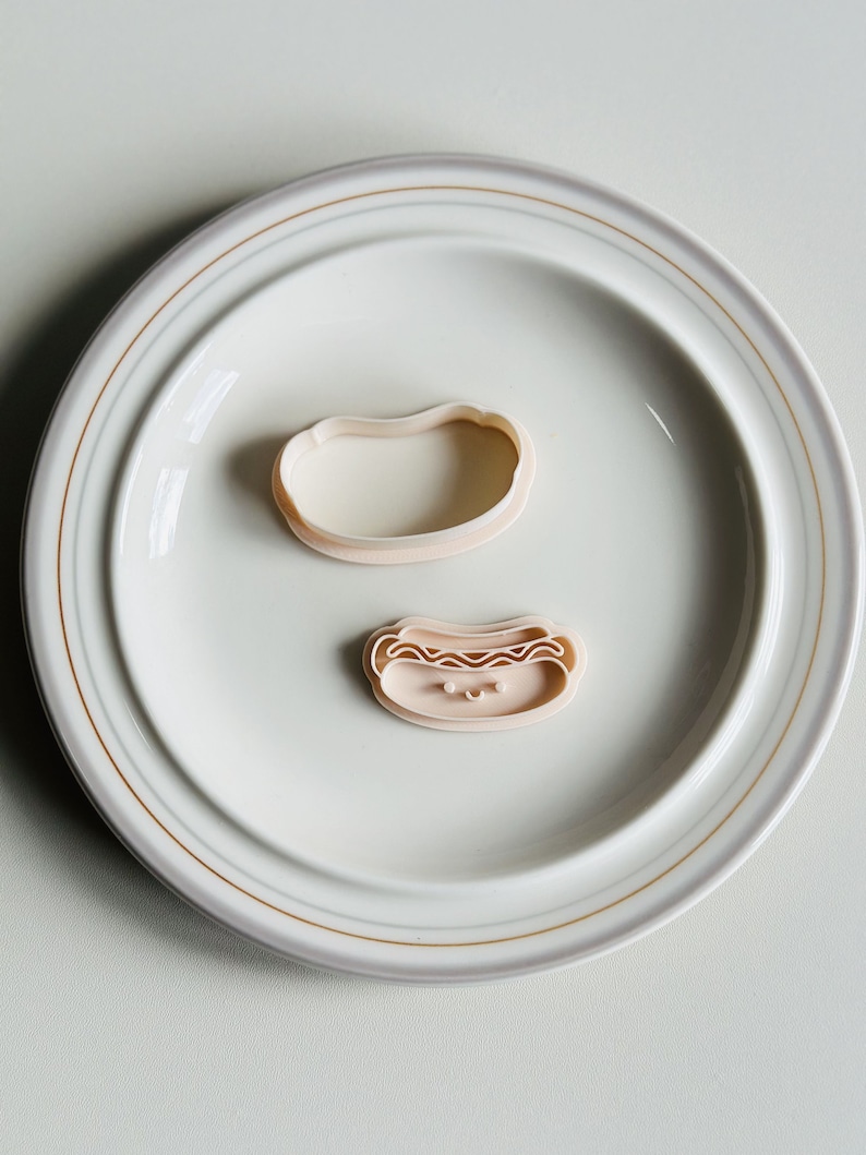 May include: Two pink plastic cookie cutters on a white plate. One cutter is shaped like a hot dog with a smiling face and the other is shaped like a bun.