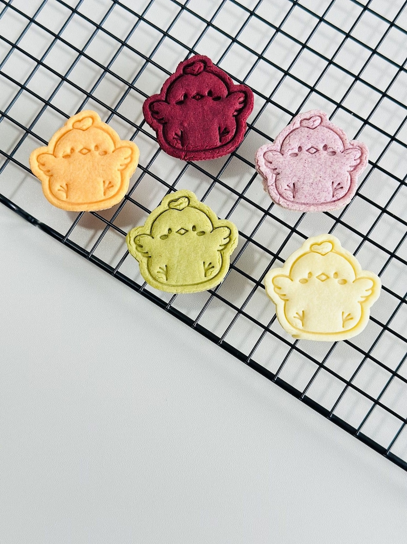 Op de afbeelding: Zes koekjes in de vorm van kuikens in verschillende kleuren op een zwart draadrek. De koekjes zijn in tinten geel, rood-paars, roze, groen en cr&egrave;me. Elk koekje heeft een gedetailleerd ontwerp van een kuiken met vleugels en een hart.