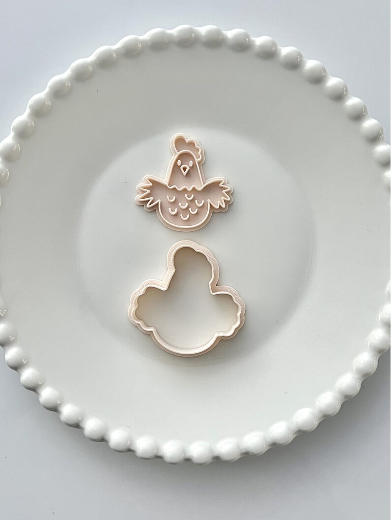 May include: Two plastic cookie cutters, one shaped like a chicken and the other shaped like a cloud, on a white plate with a beaded edge.
