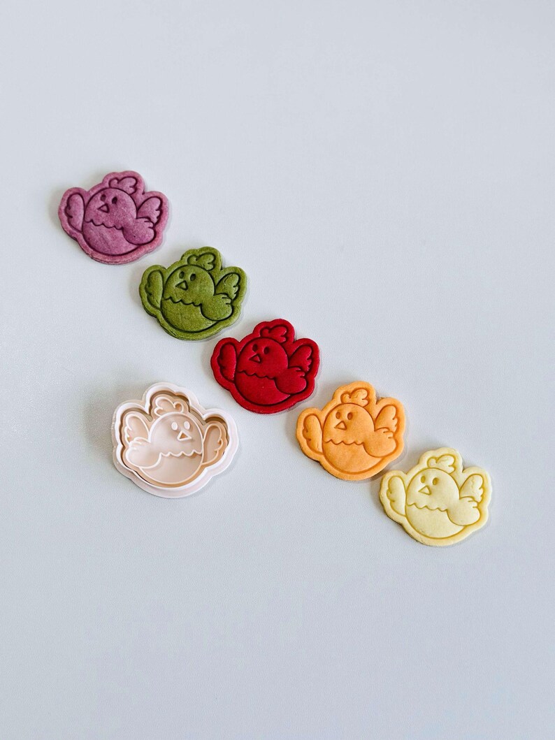 Op de afbeelding: Een verzameling koekjessnijders en koekjes in de vorm van kuikens. De koekjes zijn in de kleuren paars, groen, rood, oranje en geel. De koekjessnijder is lichtroze. Het kuikenontwerp heeft vleugels en een snavel.