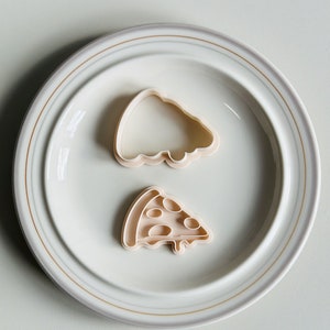 May include: Two plastic cookie cutters on a white plate. One cutter is shaped like a pizza slice and the other is shaped like a whole pizza.