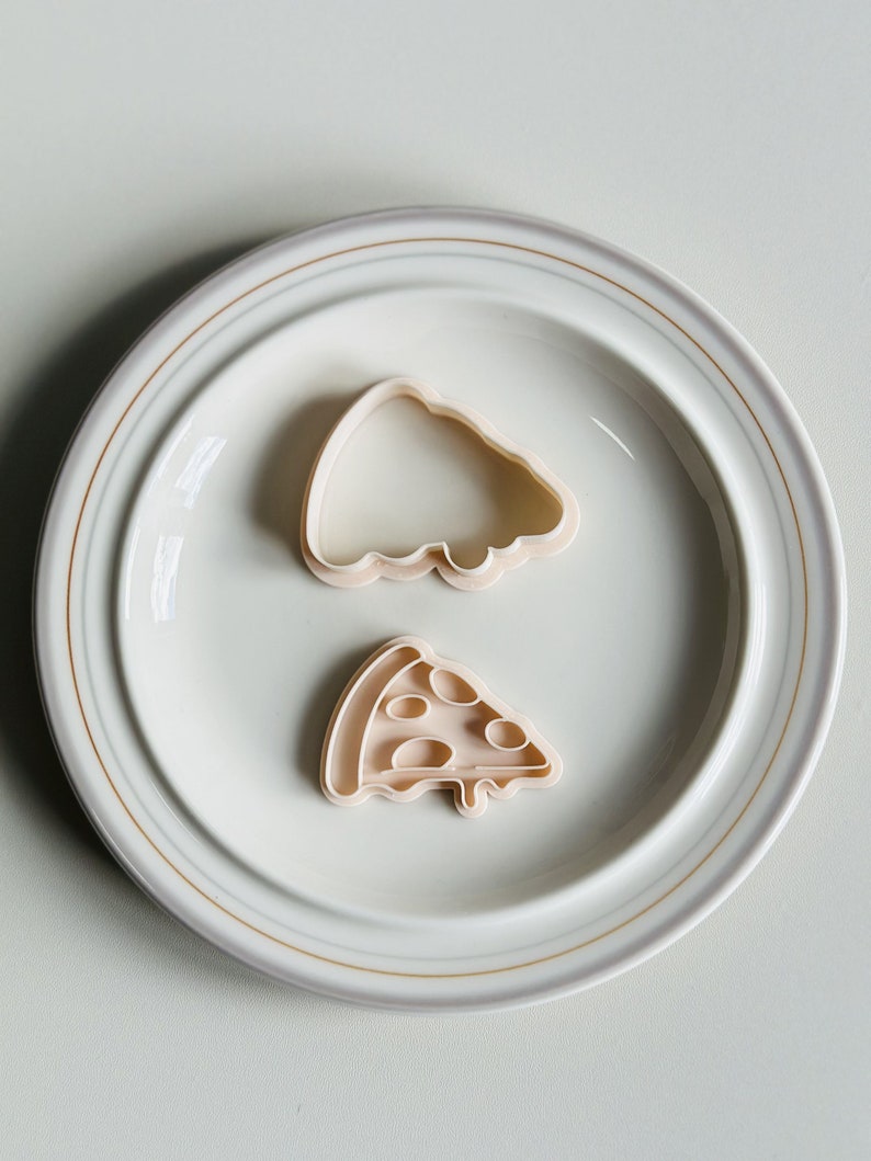 May include: Two plastic cookie cutters on a white plate. One cutter is shaped like a pizza slice and the other is shaped like a whole pizza.