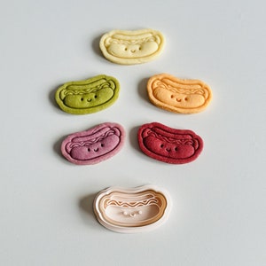 May include: Five colorful hot dog shaped cookies with happy faces and a white plastic cookie cutter in the shape of a hot dog with a happy face.