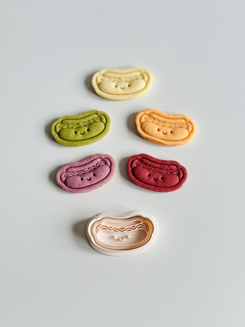 May include: Five colorful hot dog shaped cookies with happy faces and a white plastic cookie cutter in the shape of a hot dog with a happy face.
