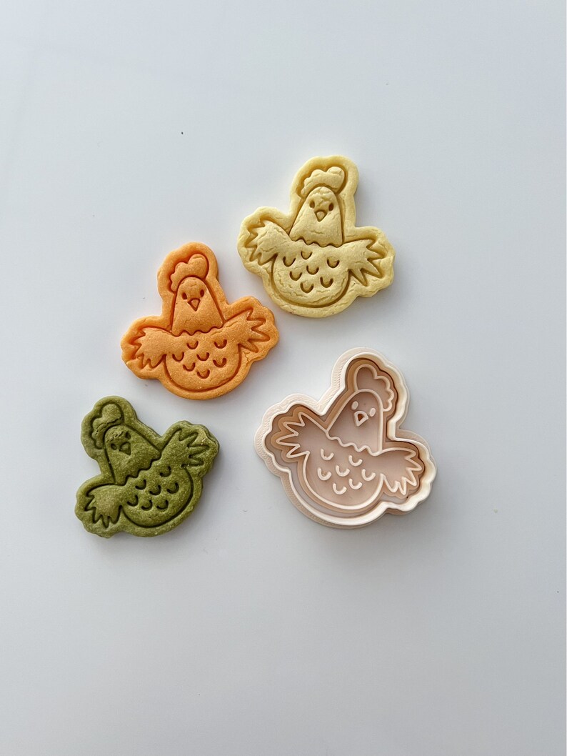 May include: Three cookie cutters in the shape of a chicken. The cutters are in different colors: yellow, orange, and green. The cutters are on a white background.