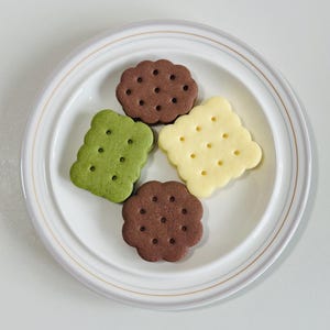 May include: Four different flavored cookies on a white plate. The cookies are green, brown, and yellow.