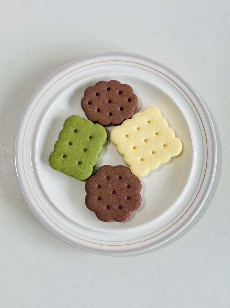 May include: Four different flavored cookies on a white plate. The cookies are green, brown, and yellow.