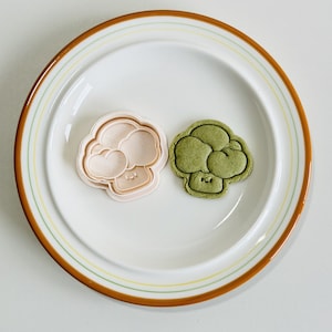 May include: Two broccoli-shaped cookie cutters, one pink and one green, on a white plate with a brown rim.