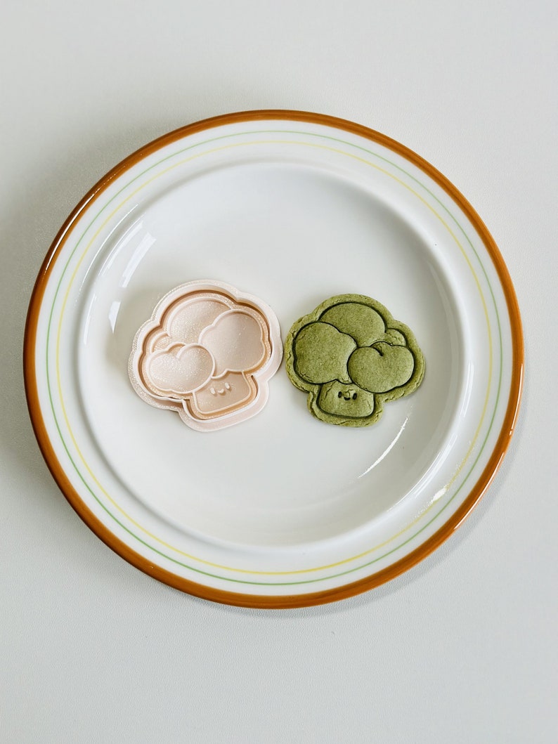 May include: Two broccoli-shaped cookie cutters, one pink and one green, on a white plate with a brown rim.