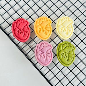 May include: Six heart-shaped cookies in different colors: red, orange, yellow, pink, green, and white. The cookies are arranged on a black wire rack.