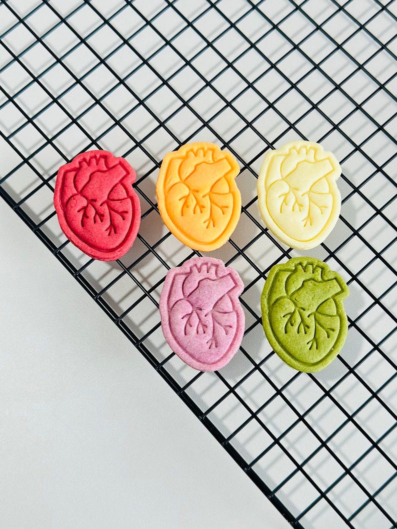 May include: Six heart-shaped cookies in different colors: red, orange, yellow, pink, green, and white. The cookies are arranged on a black wire rack.