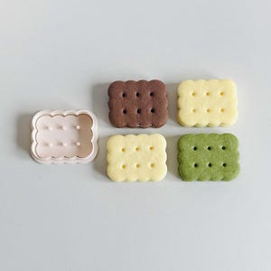 May include: Four square-shaped cookies, two with a light brown color, one with a yellow color, and one with a green color. The cookies have a pattern of small circles on the top. There is also a plastic cookie cutter in the shape of a square with a wavy edge.