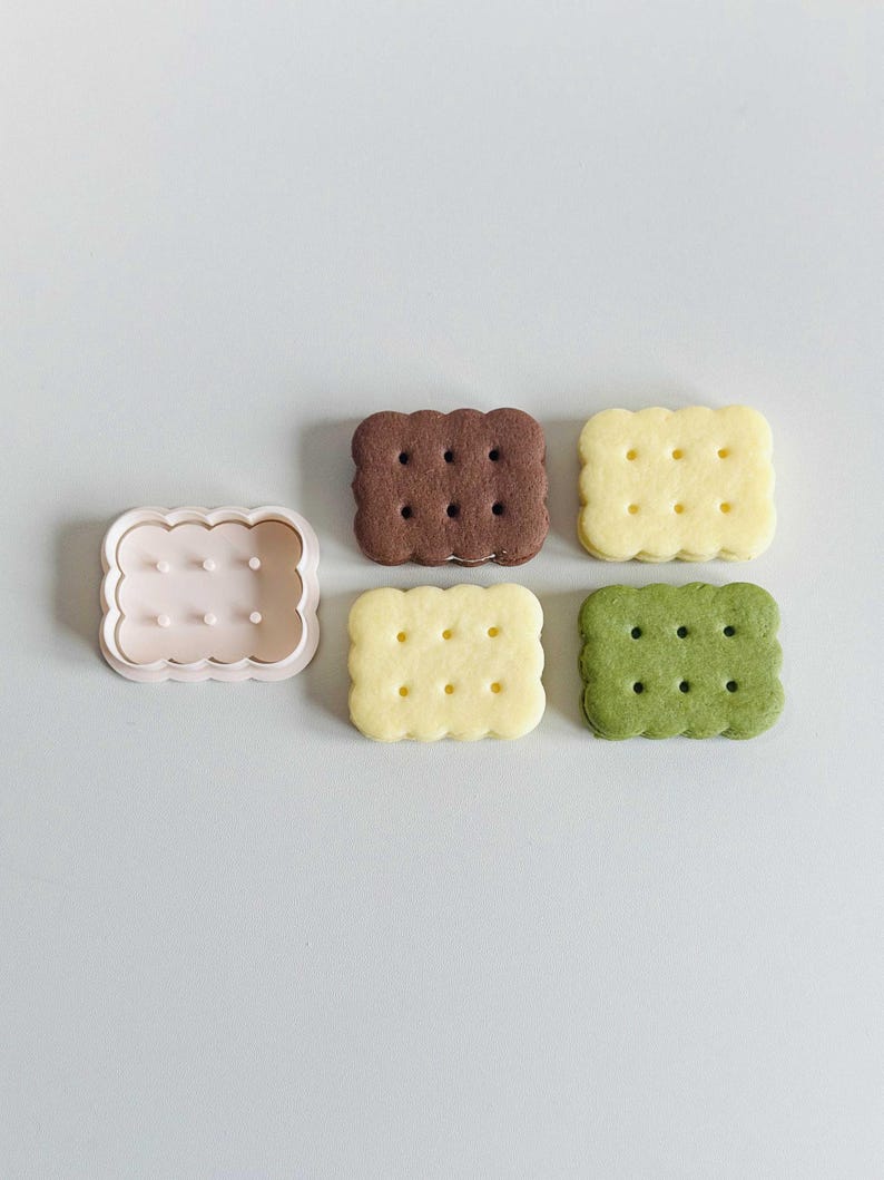 May include: Four square-shaped cookies, two with a light brown color, one with a yellow color, and one with a green color. The cookies have a pattern of small circles on the top. There is also a plastic cookie cutter in the shape of a square with a wavy edge.
