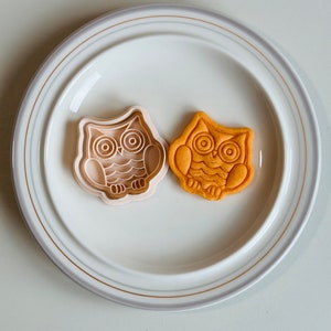 May include: A white ceramic plate with a gold rim features two owl-shaped cookie cutters. One cutter is made of white plastic and the other is a golden brown cookie.