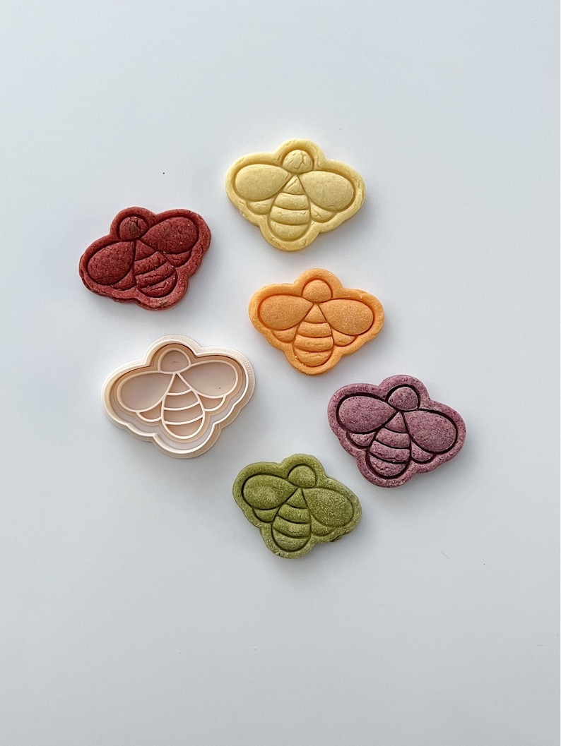 May include: A set of six bee-shaped cookie cutters, including one white plastic cutter and five colorful cookies. The cookies are yellow, orange, red, green, and purple.