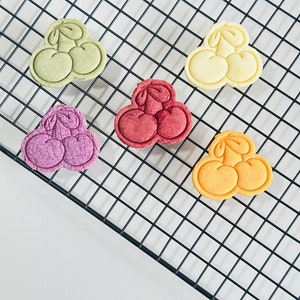 May include: Five colorful cookies shaped like cherries on a black wire cooling rack. The cookies are green, yellow, red, purple, and orange.
