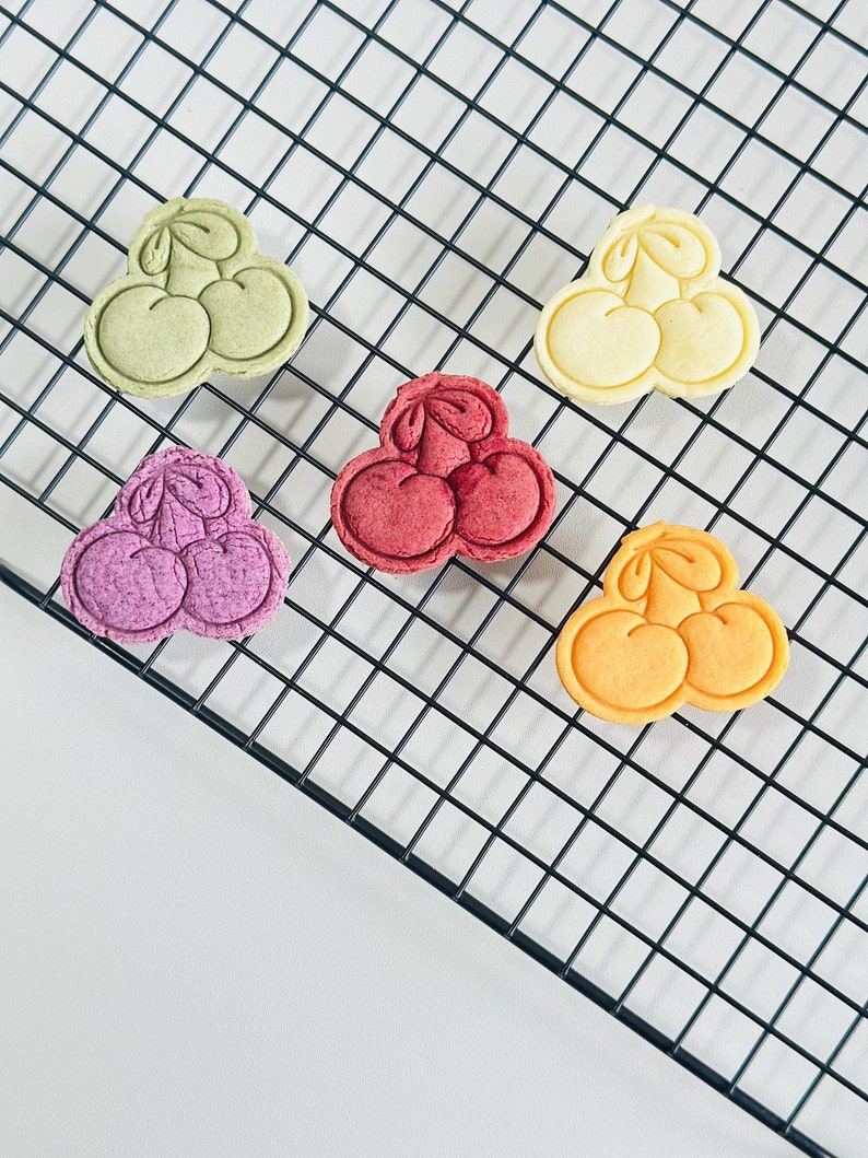 May include: Five colorful cookies shaped like cherries on a black wire cooling rack. The cookies are green, yellow, red, purple, and orange.