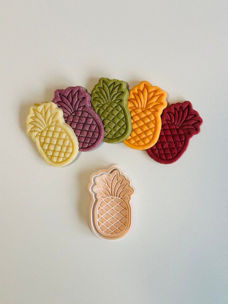 May include: Five pineapple-shaped cookie cutters in different colors: yellow, green, purple, red, and beige. The cutters are arranged in a semi-circle with the beige cutter at the bottom. A white plastic pineapple cookie cutter is in the center.