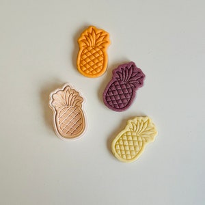 May include: Four pineapple-shaped cookie cutters in different colors: orange, purple, beige, and yellow. The cutters are on a light beige surface.