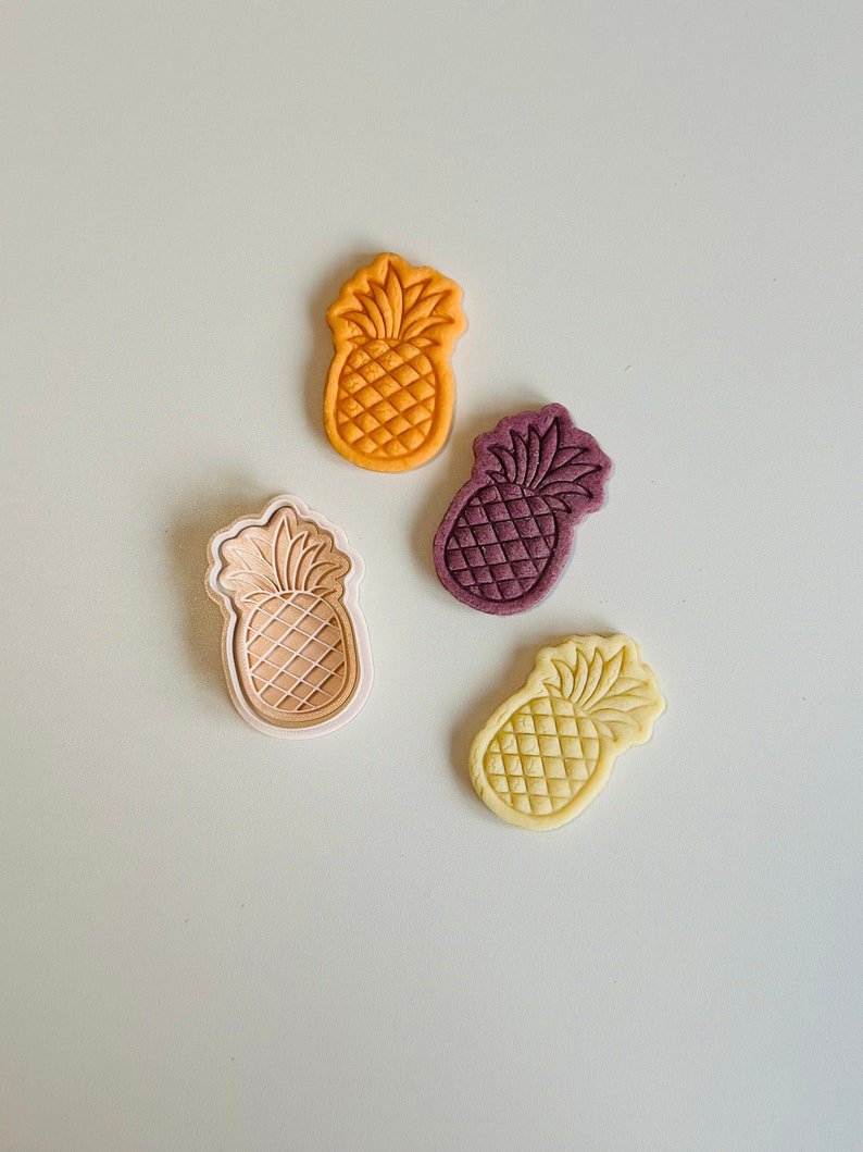 May include: Four pineapple-shaped cookie cutters in different colors: orange, purple, beige, and yellow. The cutters are on a light beige surface.