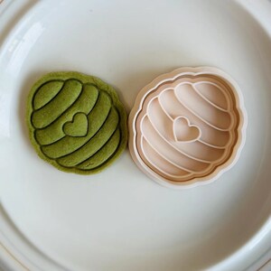 Pu&ograve; includere: Un tagliapasta verde a forma di cuore con un design a spirale. Il tagliapasta &egrave; su un piatto bianco accanto a un tagliapasta beige della stessa forma.