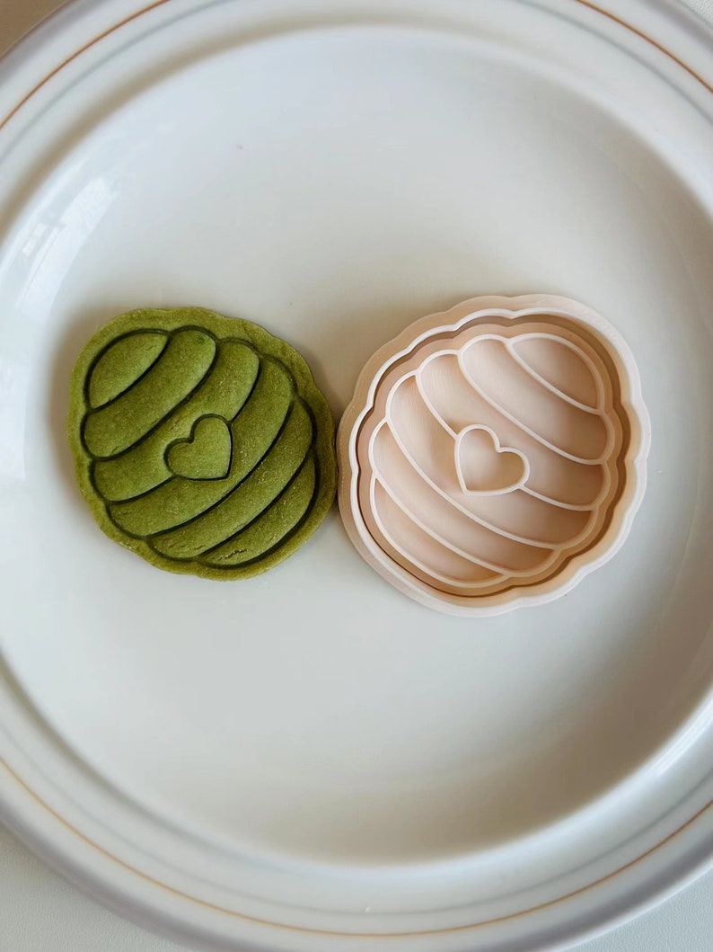Pu&ograve; includere: Un tagliapasta verde a forma di cuore con un design a spirale. Il tagliapasta &egrave; su un piatto bianco accanto a un tagliapasta beige della stessa forma.