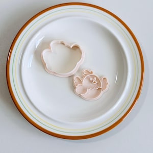 Op de afbeelding: Twee roze plastic koekjessnijders in de vorm van een vogel en een wolk liggen op een witte plaat met een bruine en gele rand. De vogeluitsteker heeft een snavel en vleugels. De wolkenuitsteker is een simpele omtrek.