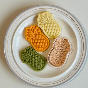 May include: Four pineapple-shaped cookies on a white plate. The cookies are green, orange, yellow, and beige.