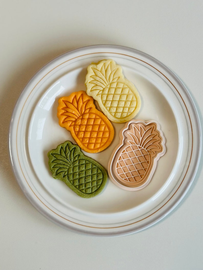 May include: Four pineapple-shaped cookies on a white plate. The cookies are green, orange, yellow, and beige.